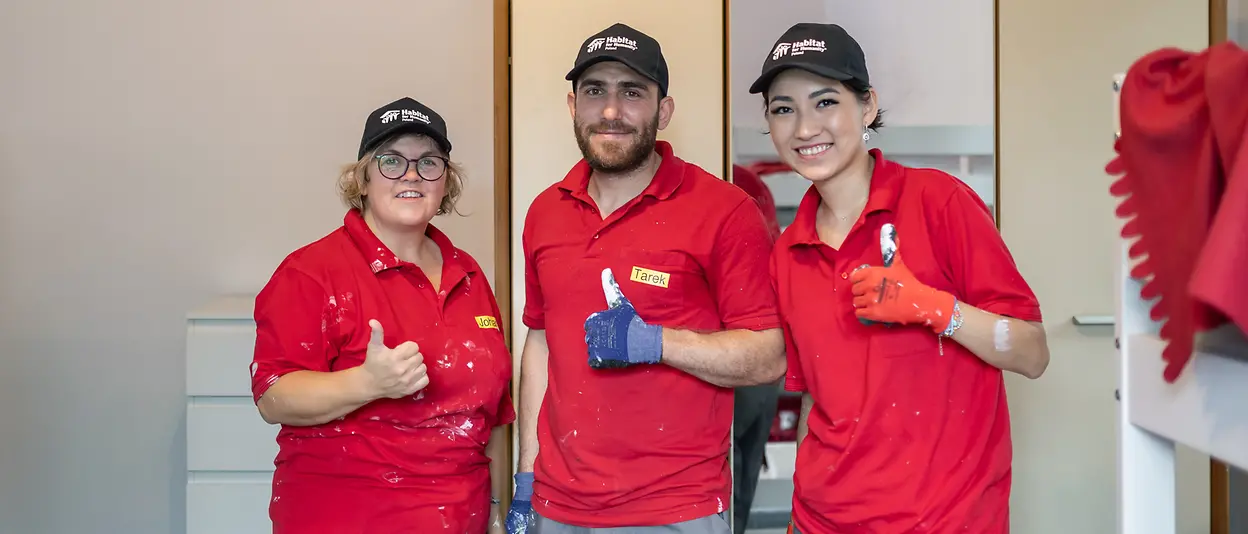 Drei Freiwillige in Arbeitskleidung und passenden T-Shirts halten ihre Daumen hoch und lächeln in die Kamera.