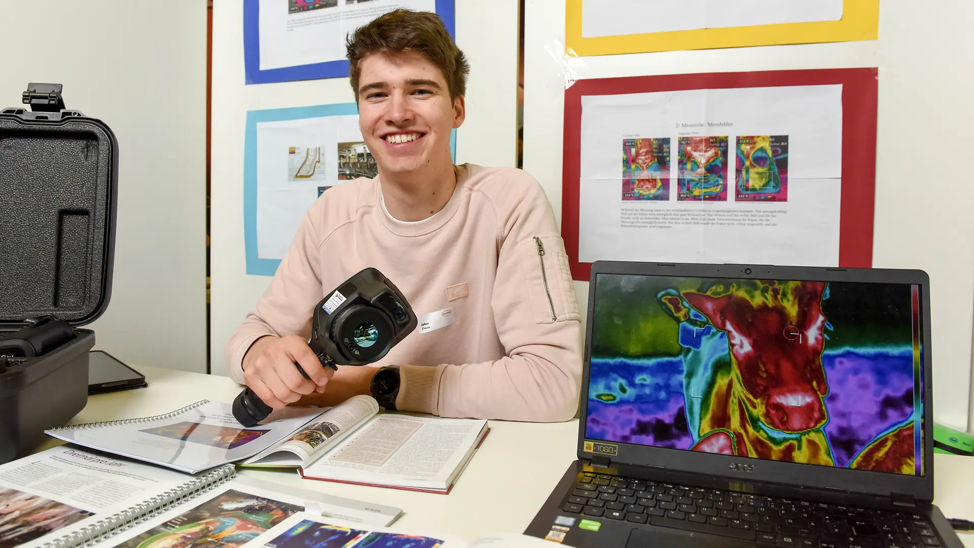 Julius Clauss sitzt lachend vor seinem Projektstand während neben ihm sein Laptop die Aufzeichnung einer Wärmebildkamera zeigt.