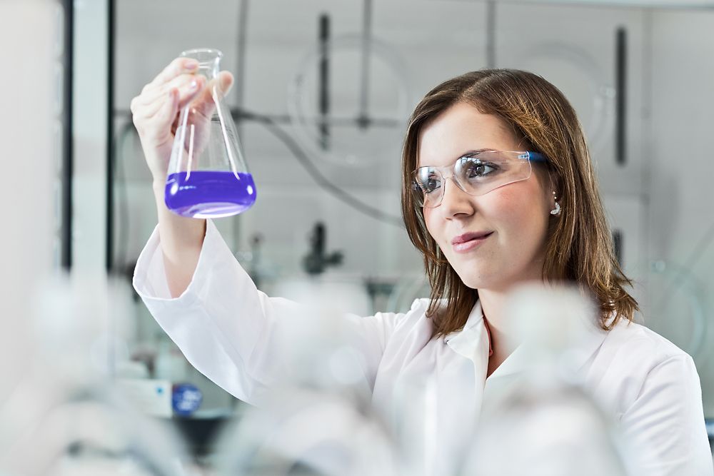 Eine Wissenschaftlerin experimentiert im Labor und trägt dabei einen Laborkittel sowie eine Sicherheitsbrille.