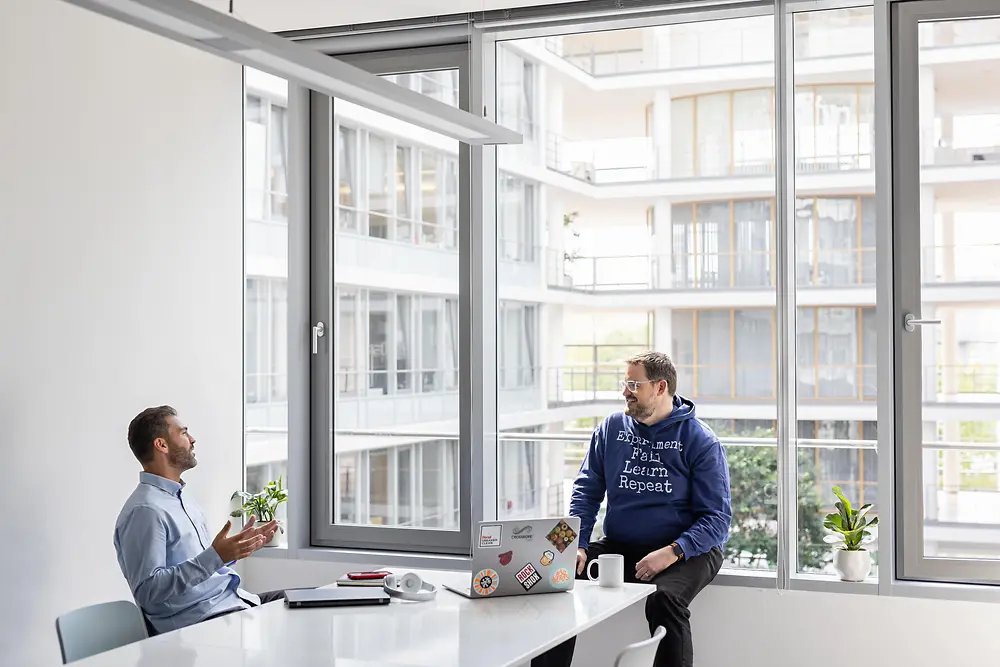 Zwei junge Männer in einem hellen Bürogebäude unterhalten sich entspannt.