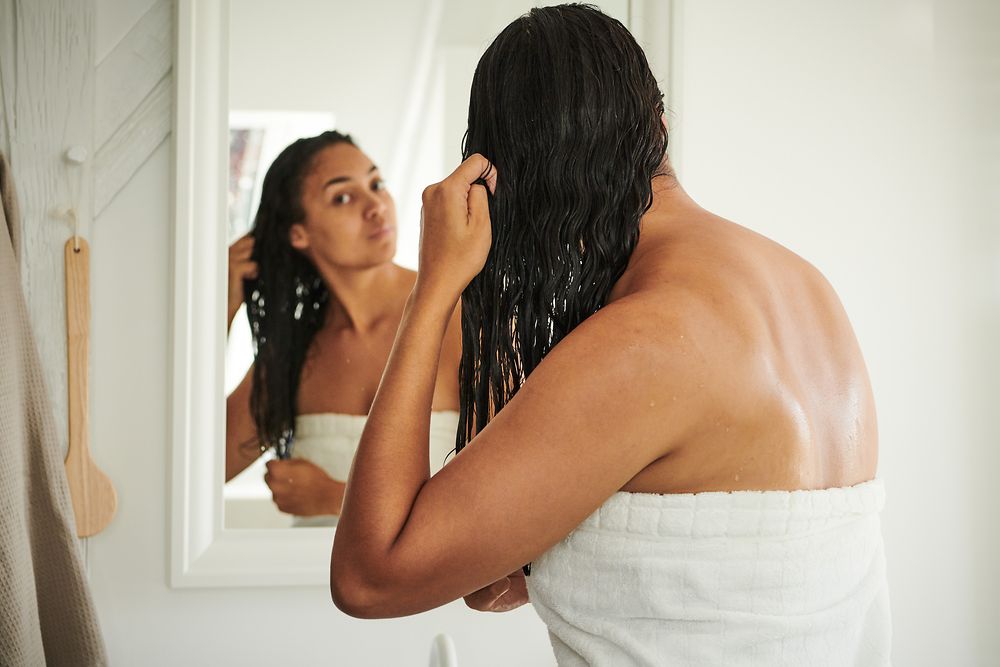 Eine Frau steht mit Badetuch und nassen Haaren vor dem Spiegel.