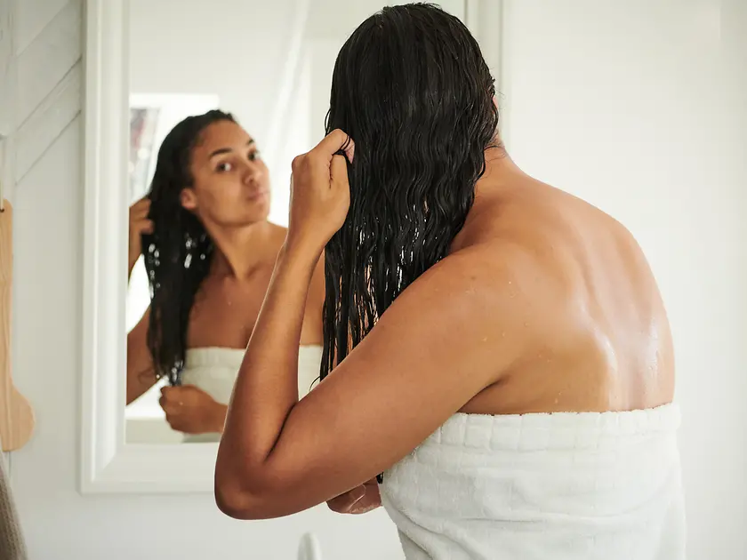 Eine Frau steht mit Badetuch und nassen Haaren vor dem Spiegel.
