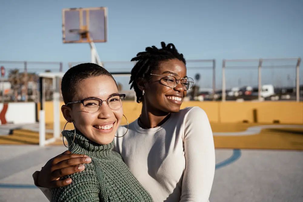 Zwei Frauen stehend lachend und umarmend auf einem Basketballplatz. 