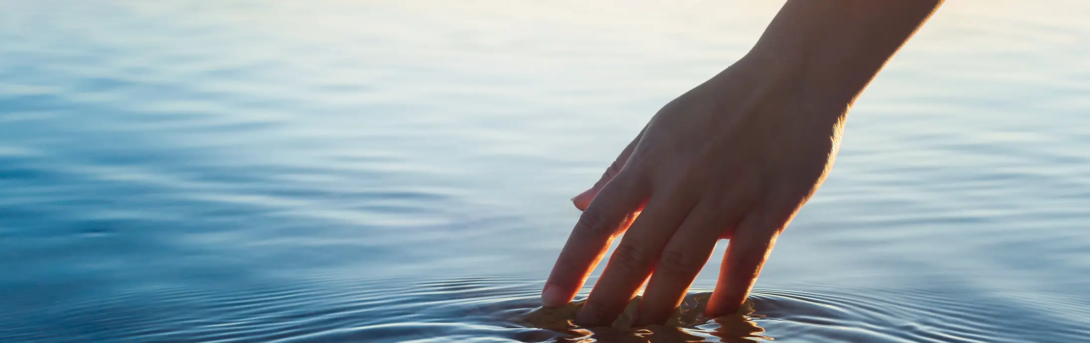 Eine Hand streicht durch eine ruhige Wasserfläche vor dem Horizont