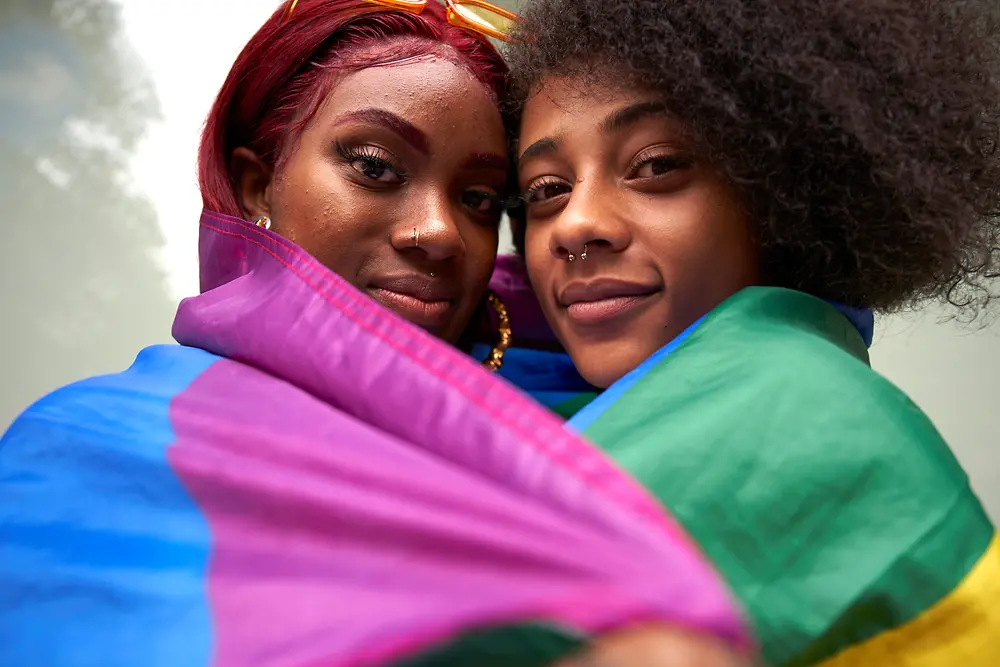 Zwei Frauen blicken lächelnd in die Kamera und haben eine Pride Flagge um sich gewickelt.