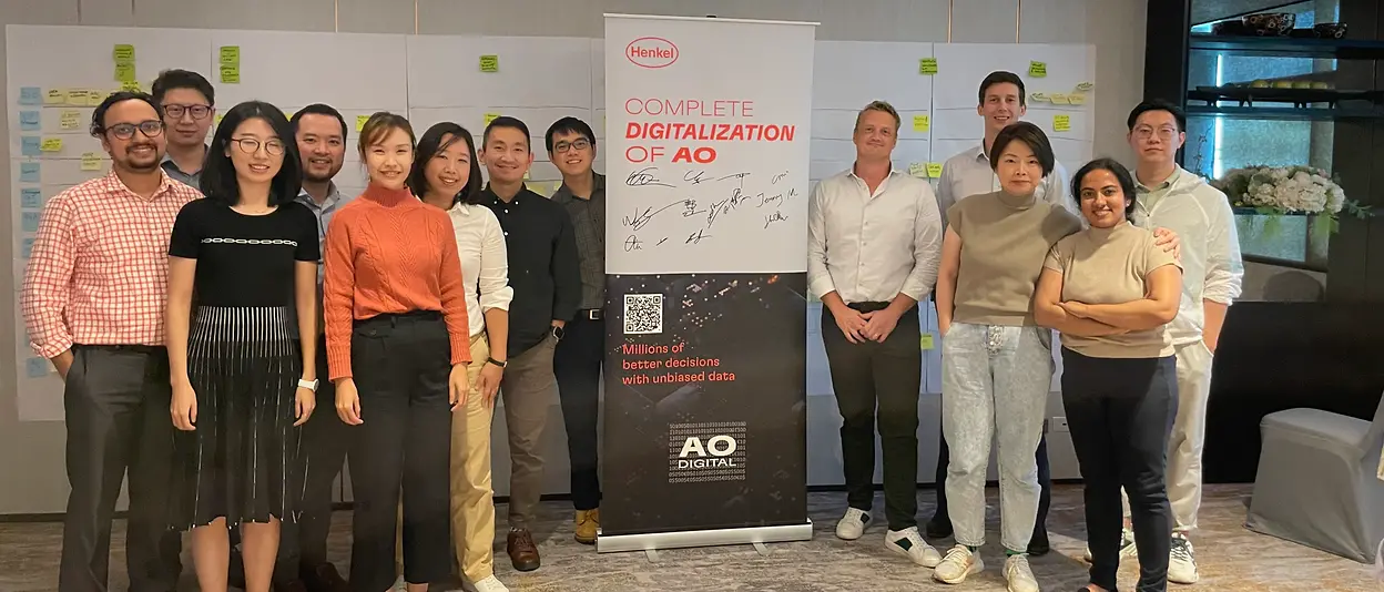 Nick und sein Team stehen zusammen in einem Besprechungsraum und posieren neben einem Roll-up mit der Aufschrift „Complete Digitalization of AO".
