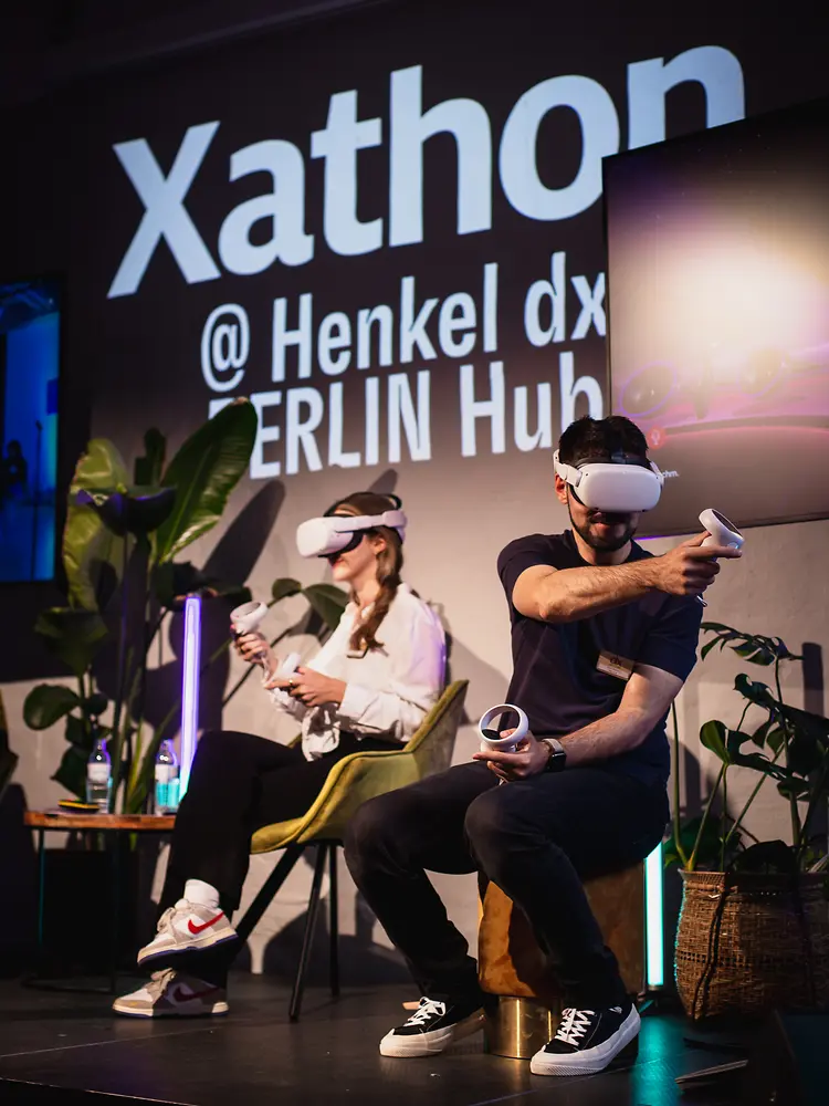 Ein Mann und eine Frau sitzen auf der Bühne, halten eine Fernbedienung und tragen eine VR-Brille.