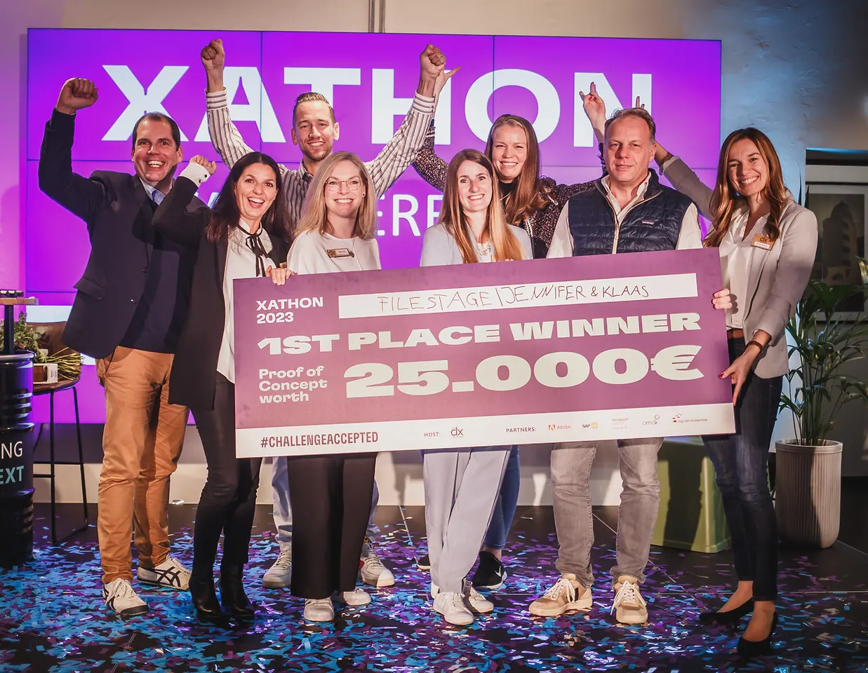 Drei Männer und fünf Frauen stehen jubelnd auf der Bühne und halten einen Scheck mit der Aufschrift „1. Platz Gewinner Proof of Concept im Wert von 25.000 Euro."