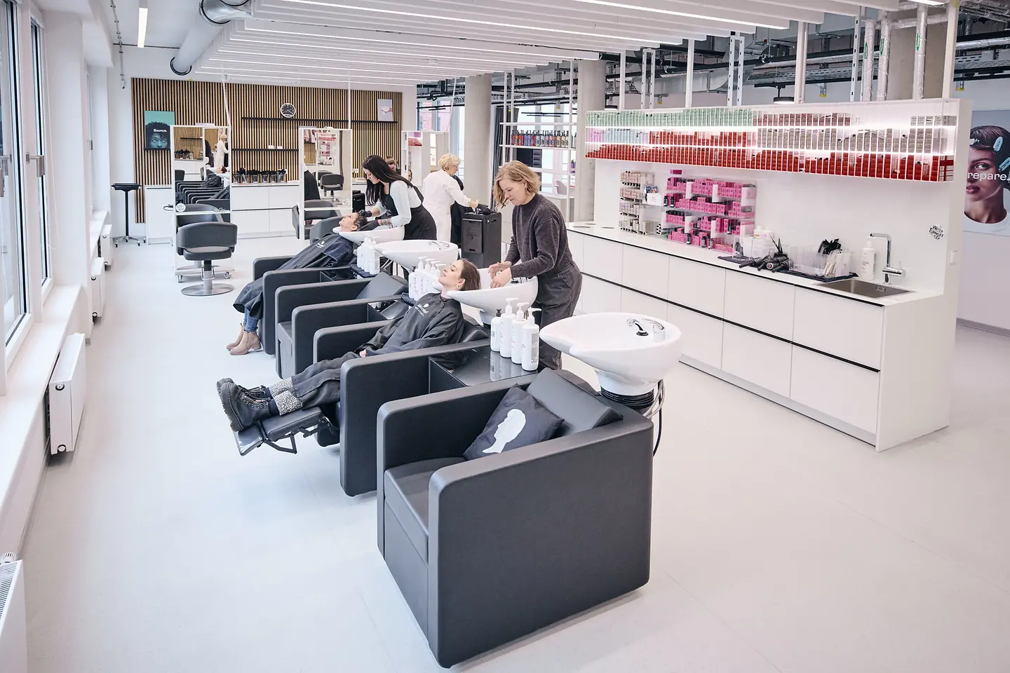 Ein heller Friseur-Salon, in dem Friseur:innen und Kund:innen zu sehen sind. 