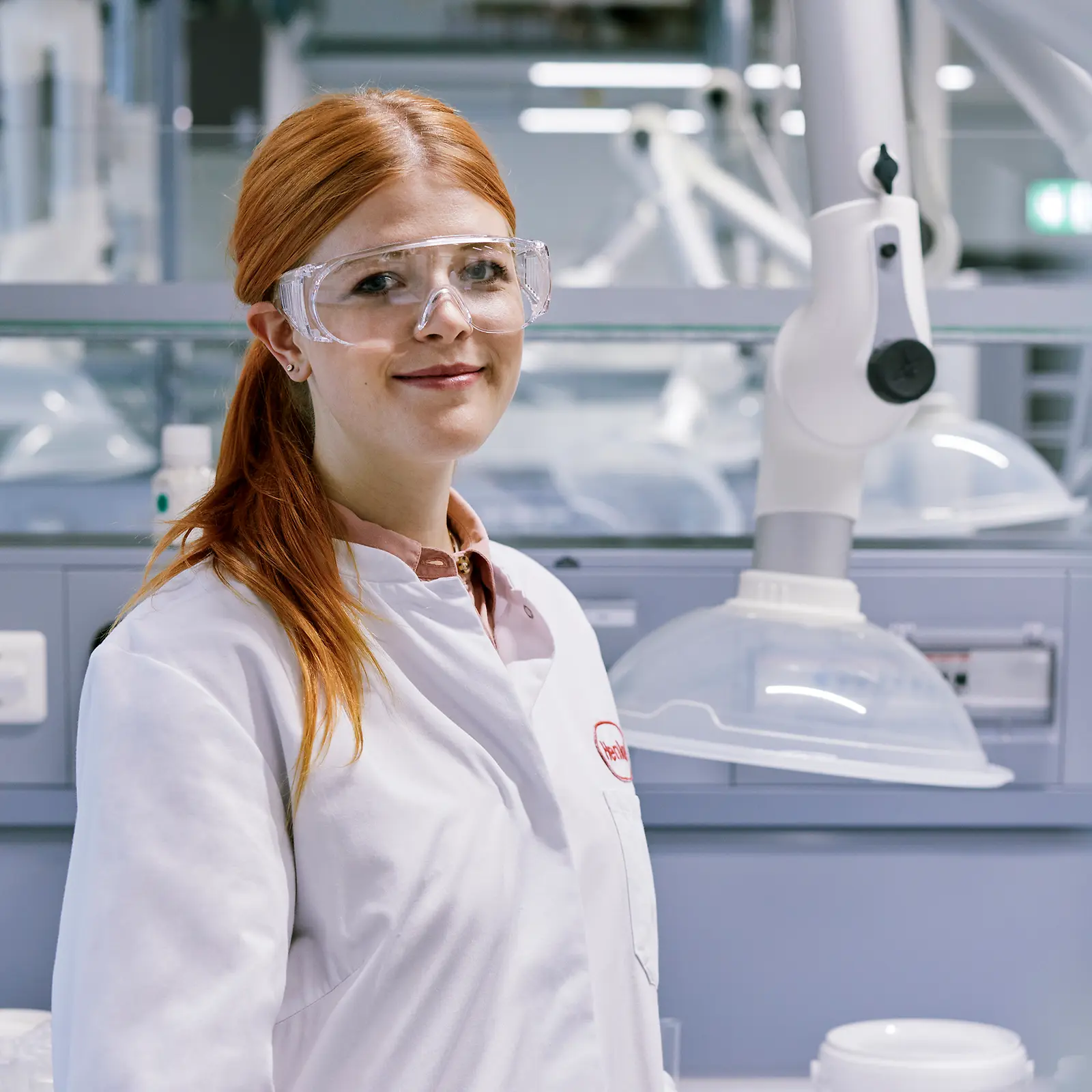 Eine rothaarige junge Frau mit Schutzbrille und weißem Laborkittel steht in einem modernen Chemielabor.