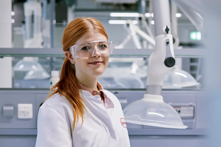 Eine rothaarige junge Frau mit Schutzbrille und weißem Laborkittel steht in einem modernen Chemielabor.