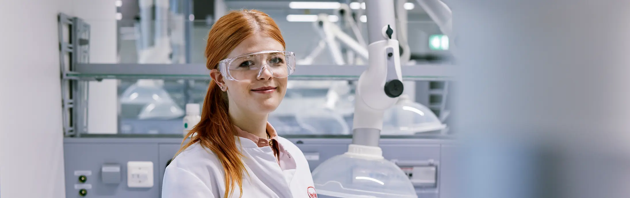 Eine rothaarige junge Frau mit Schutzbrille und weißem Laborkittel steht in einem modernen Chemielabor.