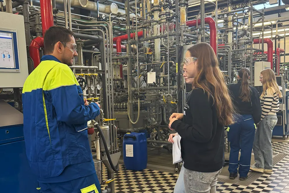 Vier Mädchen stehen mit Schutzbrillen in der Produktionstechnik vor vielen Rohren und lassen sich etwas von einem Auszubildenden erklären. 