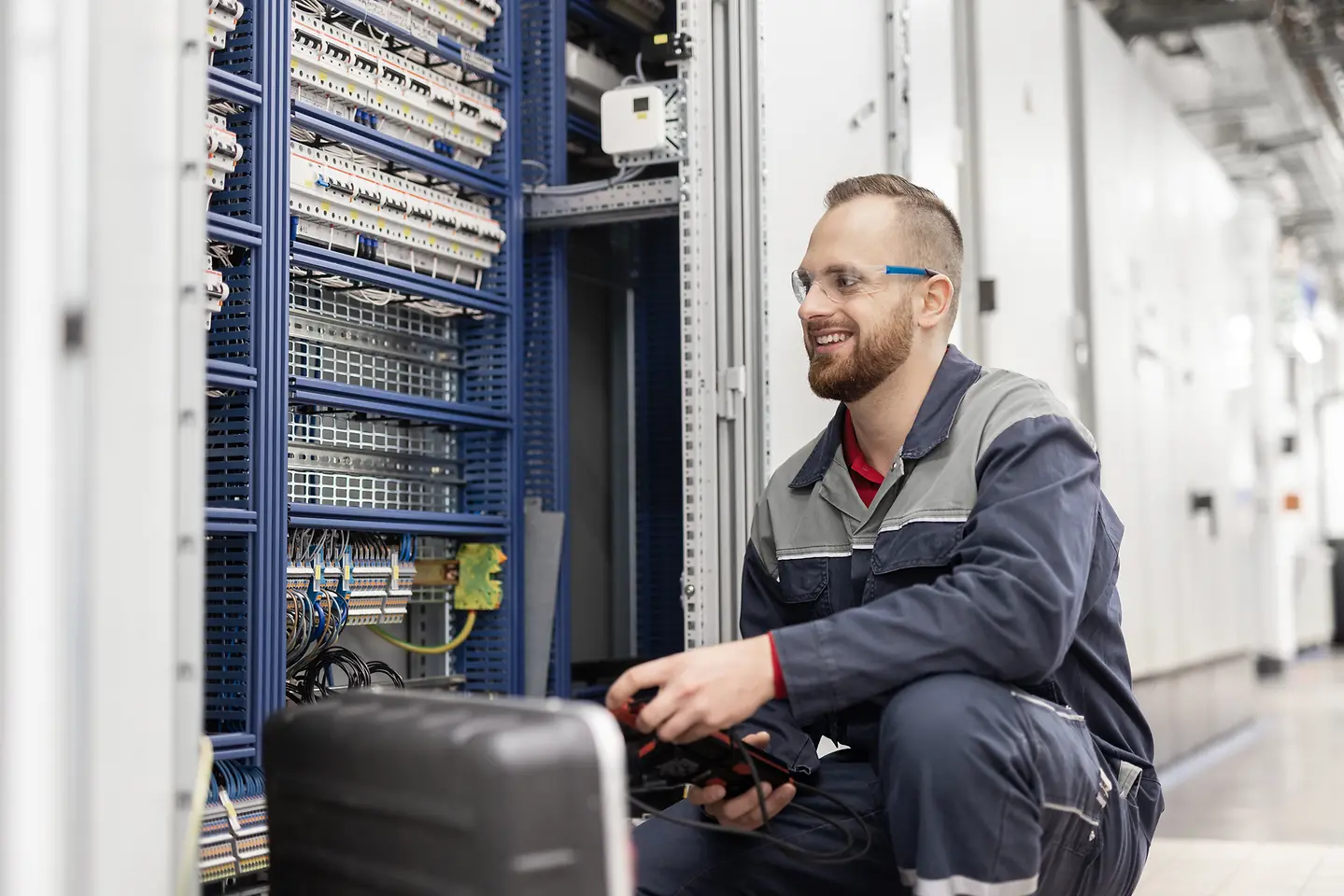 Ein Auszubildender zum Elektroniker im grauen Overall prüft einen Schaltkasten. 