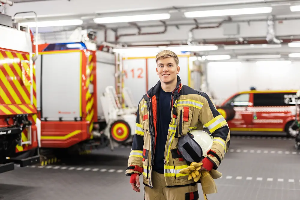 Ein Auszubildender zum Werkfeuerwehrmann steht in Einsatzkleidung in der Fahrzeughalle, in der verschiedene Feuerwehrautos parken. 