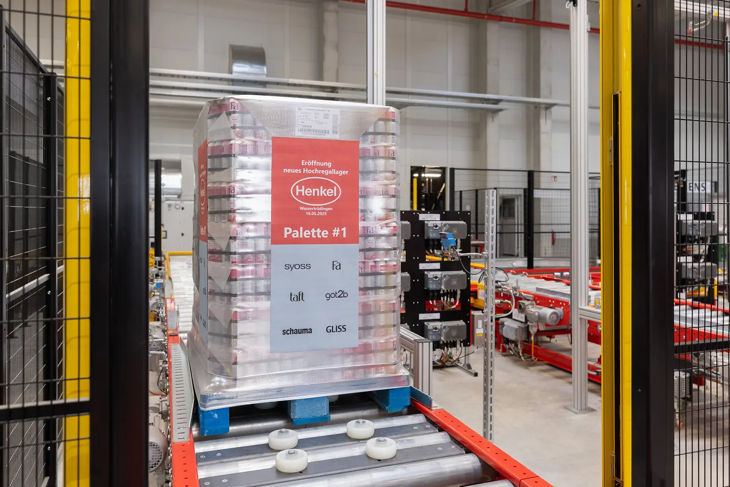 Eine Palette mit einem großen Aufkleber mit dem Henkel-Logo und der Aufschrift „Eröffnung neues Hochregallager Wassertrüdingen 16.05.2025 – Palette #1“ fährt über ein Förderband ins Lager.