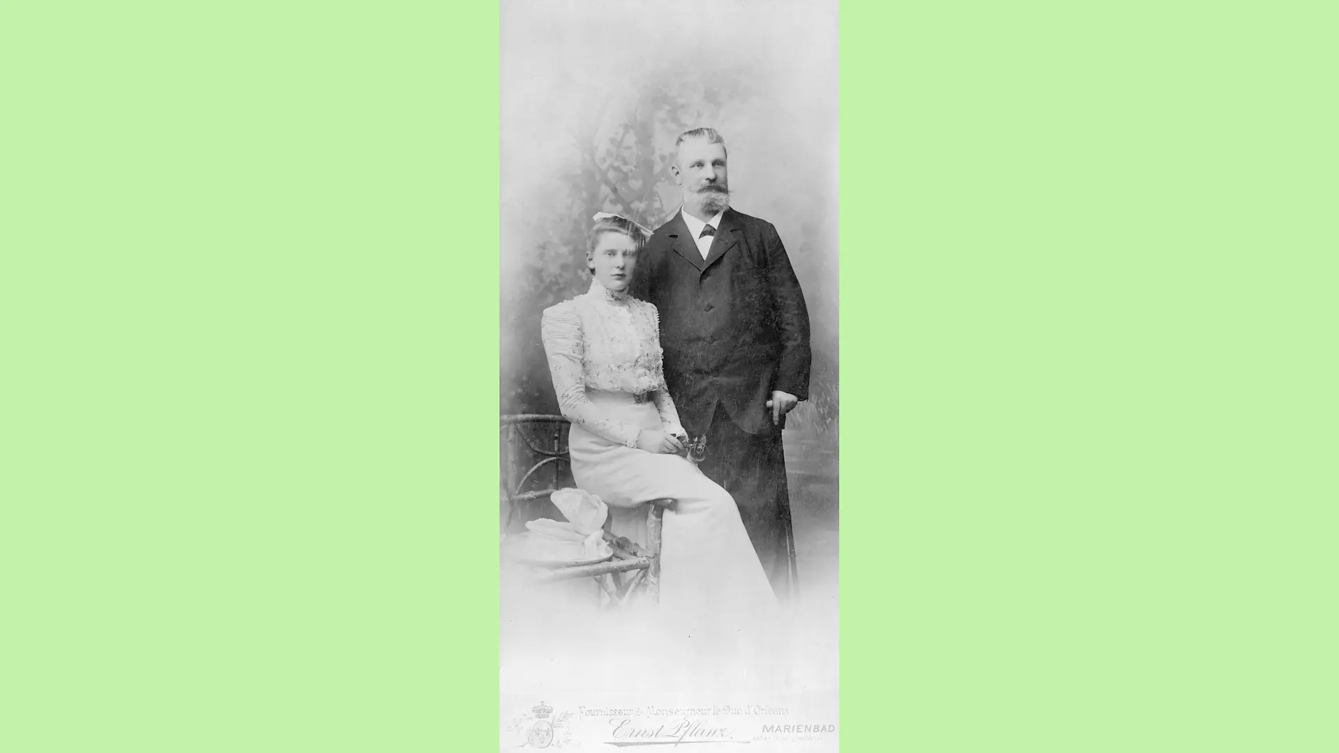 Emmy Lüps in ihrem Hochzeitskleid seitlich auf einer Stuhllehne sitzend. Neben ihr steht ihr Vater Fritz Henkel, der seinen Blick in die Ferne richtet und einen Stock in seiner linken Hand hält.