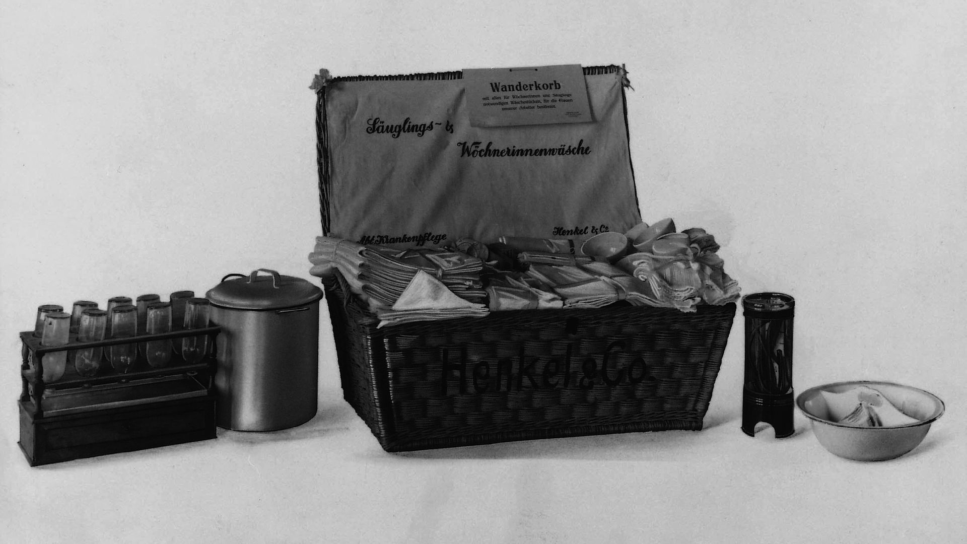 Traveling basket with baby equipment for women who recently gave birth. The company name Henkel & Cie is painted on the outside of the basket. Sterilization utensils are placed next to the basket. Textiles peek out of the open basket.