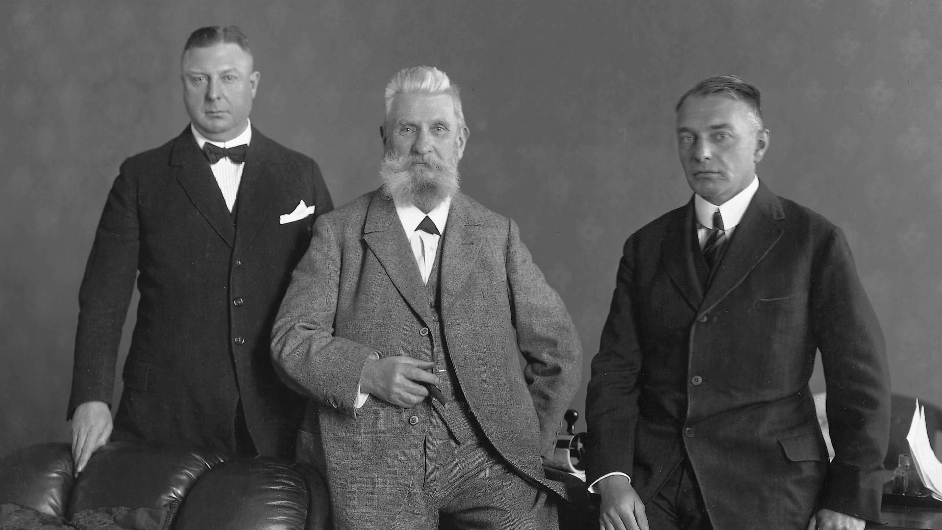 Schwarz-weiß Aufnahme von Fritz Henkel und seinen Söhnen Fritz junior und Hugo. Fritz Henkel sen. ist halb sitzend/lehnend in der Mitte platziert. Fritz jun. steht an seiner rechten Seite, seine Beine werden von einem Ledersofa verdeckt. Hugo steht an einen Tisch gelehnt auf der linken Seite seines Vaters.