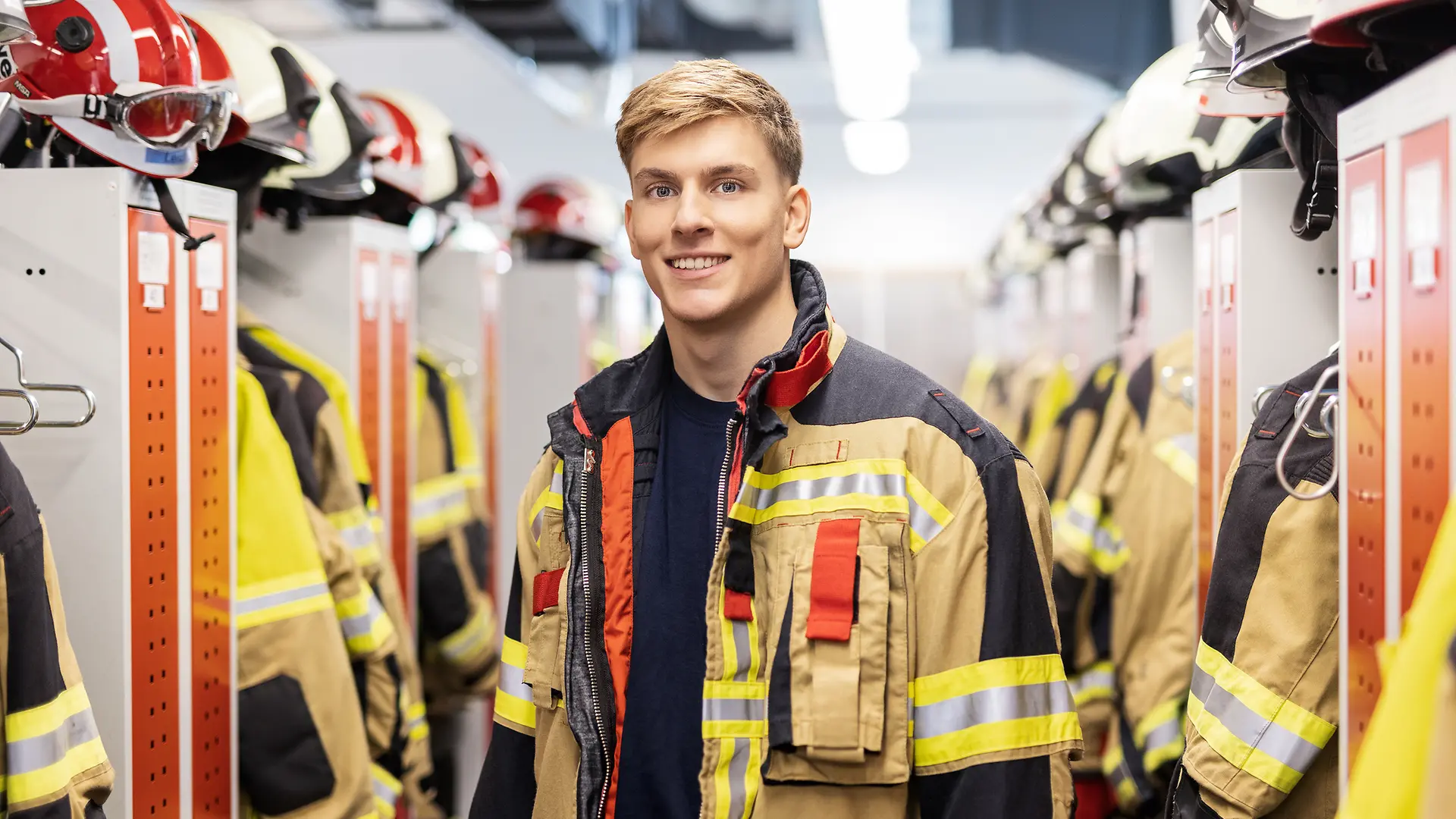 Ein Foto von einem lächelnden, jungen Mann, der eine Feuerwehruniform trägt und in einer Umkleide der Feuerwehr steht.