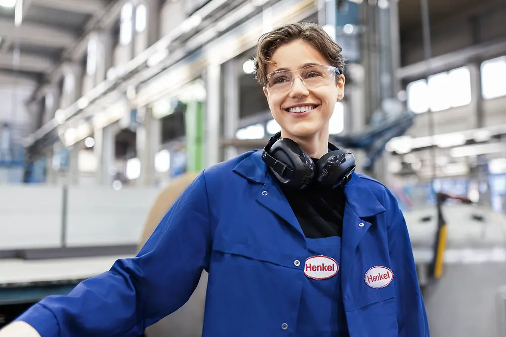 Das Foto zeigt eine lächelnde, junge Frau, die sich in einer Produktionshalle befindet und blaue Arbeitskleidung von Henkel trägt.