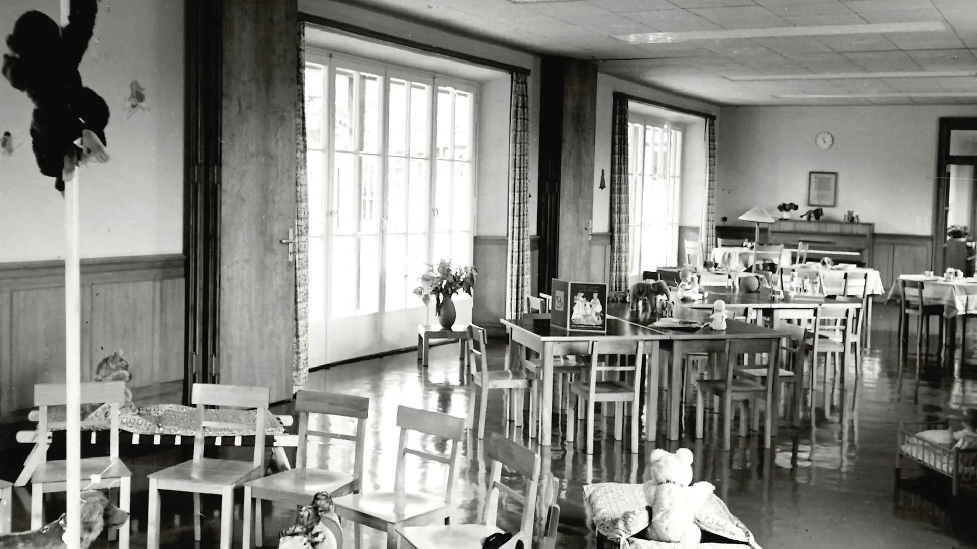 Ein Schwarz-Weiß-Foto eines geräumigen Zimmers mit hohen Fenstern. Zu sehen sind mehrere von Stühlen umgebene Tische in Kindergröße, die mit Plüschtieren und anderem Spielzeug besetzt sind.