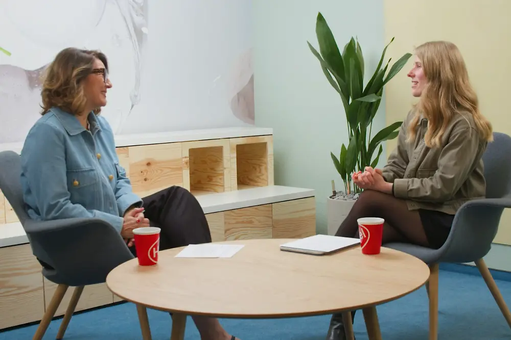 Sylvie und Leonie sitzen sich in einem modernen, hellen Raum gegenüber. Beide sitzen auf grauen Stühlen an einem runden Holztisch. Auf dem Tisch stehen zwei rote Kaffeetassen. Im Hintergrund ist eine Pflanze und ein Regal mit offenen Fächern zu sehen.