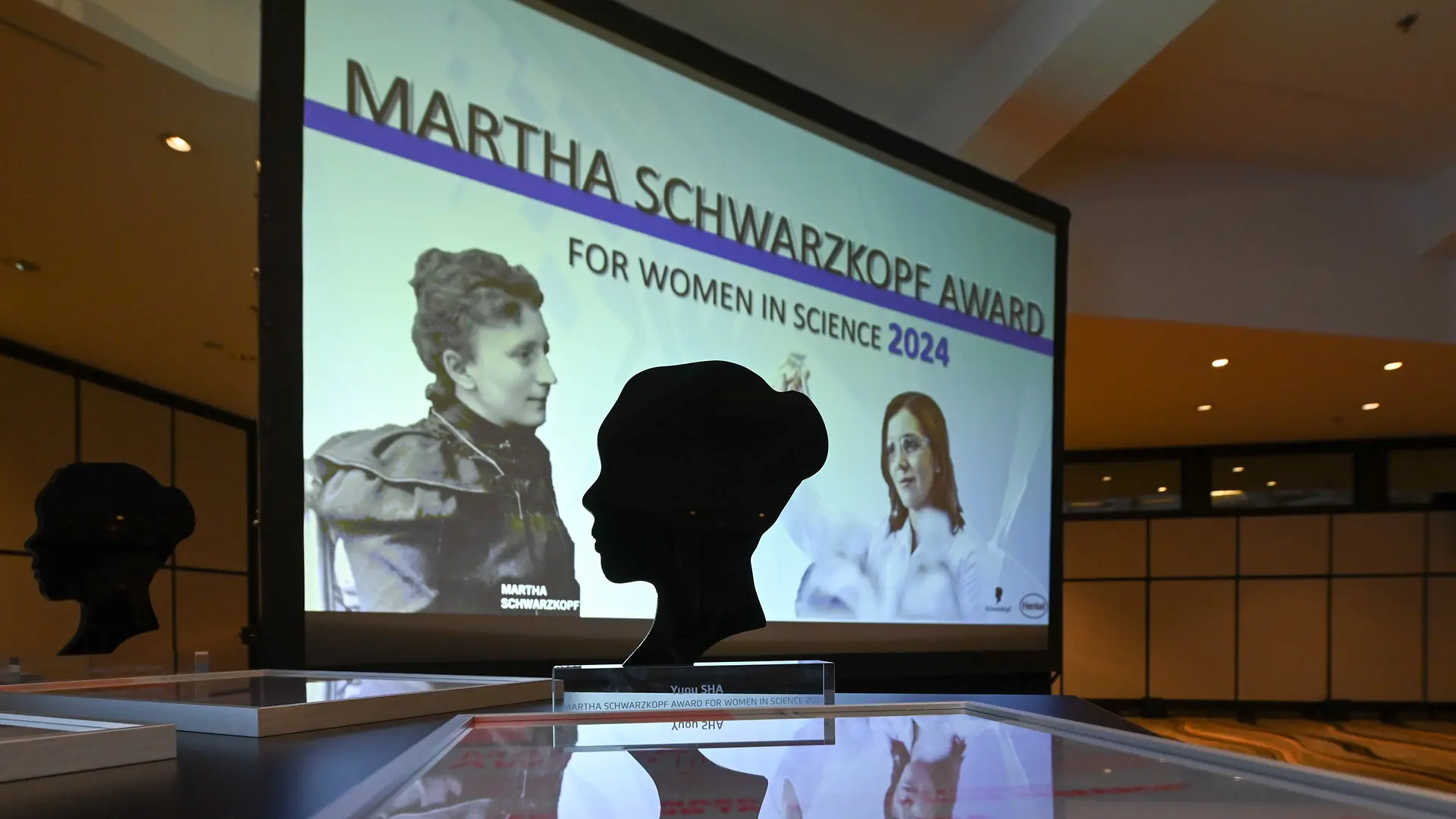 Das Bild zeigt den Martha Schwarzkopf Award auf einem Tisch. Im Hintergrund ist eine große Leinwand zu sehen, auf dem der Schriftzug „Martha Schwarzkopf Award for Women in Science 2024“ angezeigt wird. Die Szene vermittelt den feierlichen Rahmen einer Preisverleihung.