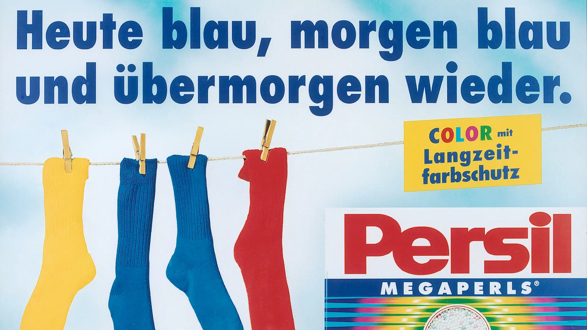 Eine Werbeanzeige für Persil Color. Eine Wäscheleine mit vier bunten Socken, darunter zwei blauen, hängt im Hintergrund des Bildes. In der rechten Ecke ist eine Packung Persil Color abgebildet und darüber steht der Satz „Color mit Langzeit-Farbschutz“. Über der Anzeige steht der Satz „Heute blau, morgen blau und übermorgen wieder“.