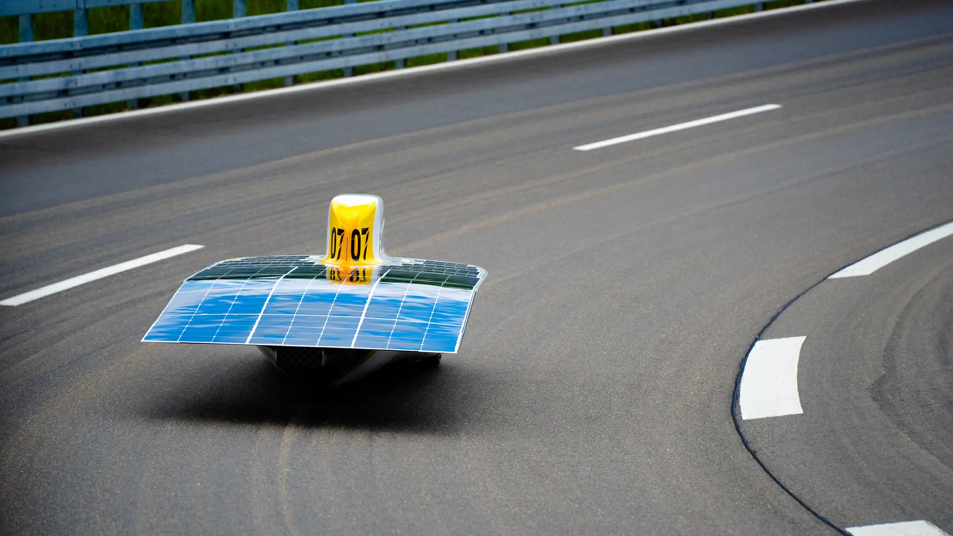 Der Sonnenwagen ist von hinten zu sehen, während er auf einer asphaltierten Straße fährt. Das Solardeck mit blauen Solarpaneelen ist deutlich sichtbar. 