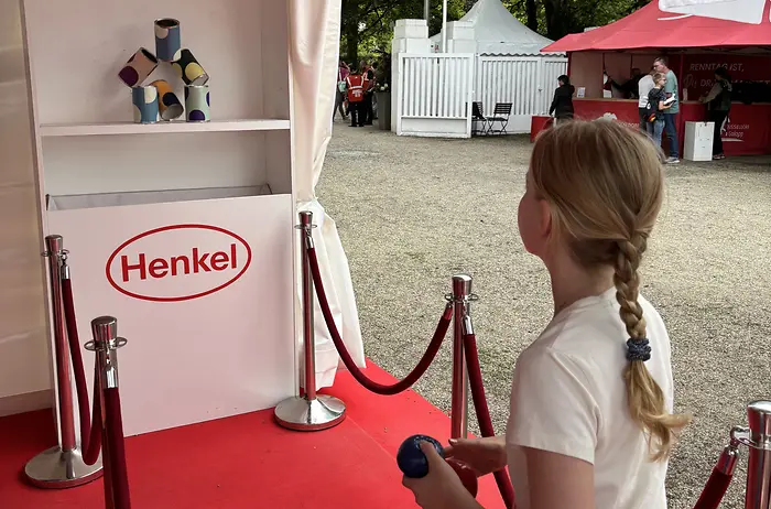 Ein Mädchen mit geflochtenen blonden Haaren von hinten gesehen, das auf einem roten Teppich in einem Pavillon mit „Fritz Henkel Stiftung“ und „Henkel“ Logos steht und eine blaue Kugel auf einen Stapel bunter Dosen wirft; im Hintergrund Bäume und weiße Zelte eines Außengeländes.