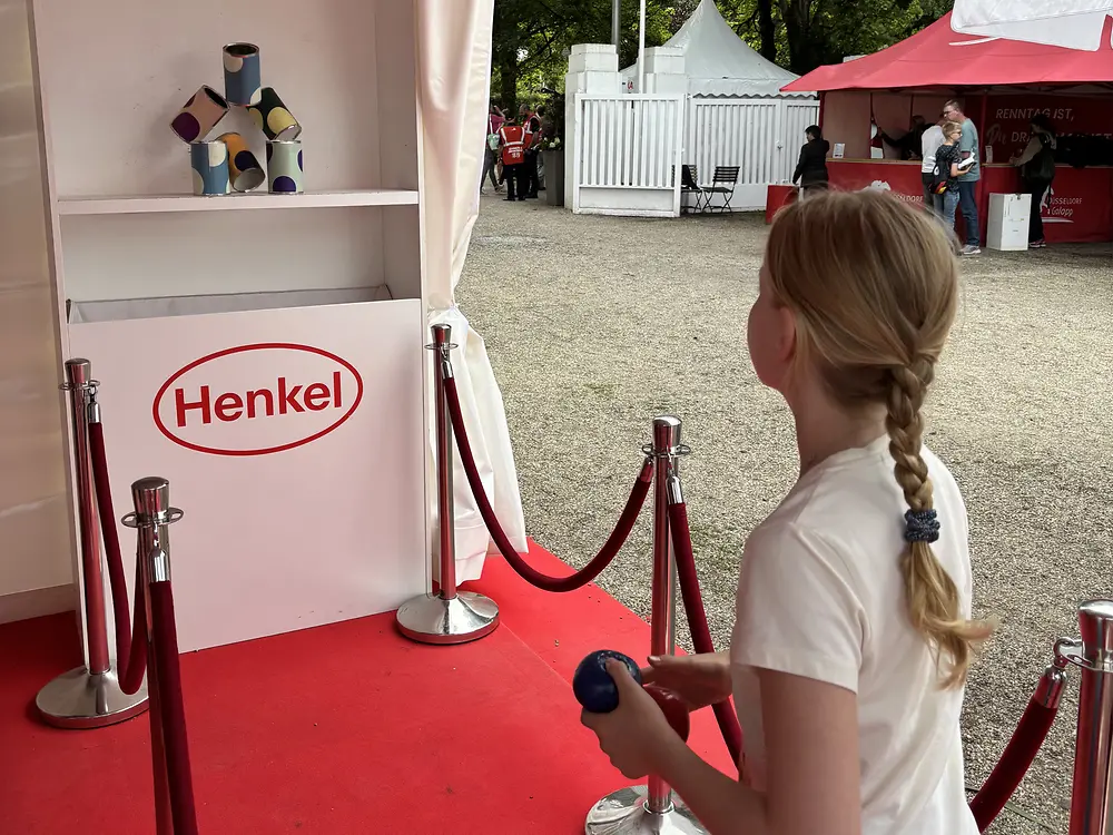 Ein Mädchen mit geflochtenen blonden Haaren von hinten gesehen, das auf einem roten Teppich in einem Pavillon mit „Fritz Henkel Stiftung“ und „Henkel“ Logos steht und eine blaue Kugel auf einen Stapel bunter Dosen wirft; im Hintergrund Bäume und weiße Zelte eines Außengeländes.