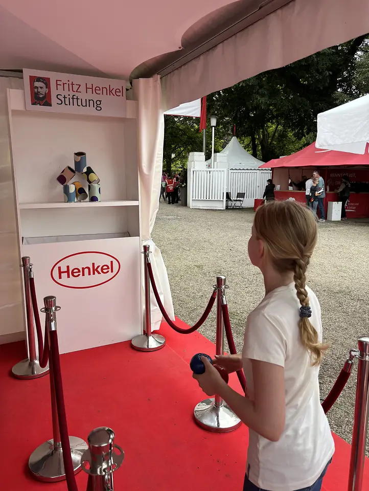Ein Mädchen mit geflochtenen blonden Haaren von hinten gesehen, das auf einem roten Teppich in einem Pavillon mit „Fritz Henkel Stiftung“ und „Henkel“ Logos steht und eine blaue Kugel auf einen Stapel bunter Dosen wirft; im Hintergrund Bäume und weiße Zelte eines Außengeländes.