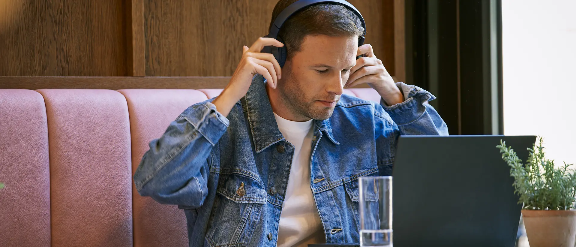 Ein Mann mit einer Jeansjacke und Kopfhörern sitzt an einem Tisch in einem Café und arbeitet an einem Laptop. Auf dem Tisch stehen ein Glas Wasser und eine Topfpflanze.
