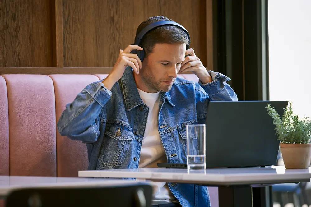 Ein Mann mit einer Jeansjacke und Kopfhörern sitzt an einem Tisch in einem Café und arbeitet an einem Laptop. Auf dem Tisch stehen ein Glas Wasser und eine Topfpflanze.