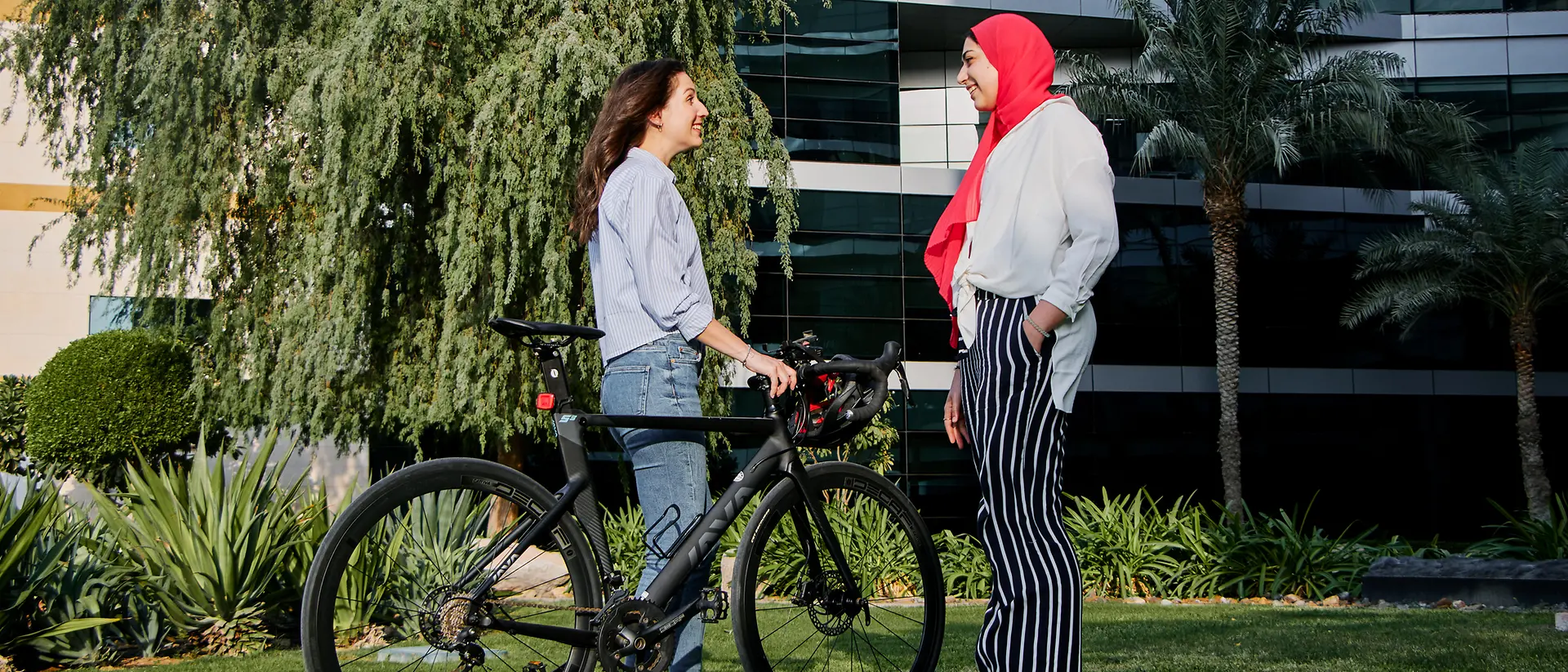 Ein Foto von zwei Frauen, die gemeinsam in einem begrünten Bereich vor einem Bürogebäude stehen – eine von ihnen hält ihr Fahrrad und ihren Helm.