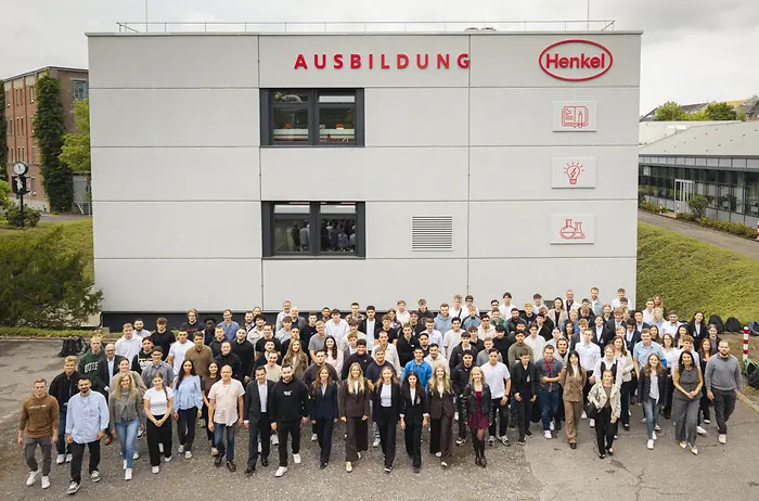 Eine große Gruppe neuer Auszubildender und dual Studierender steht vor dem Henkel-Ausbildungsgebäude in Düsseldorf zum Gruppenfoto. Über dem Gebäude ist das rote Henkel-Logo sowie die Aufschrift „Ausbildung“ zu sehen.