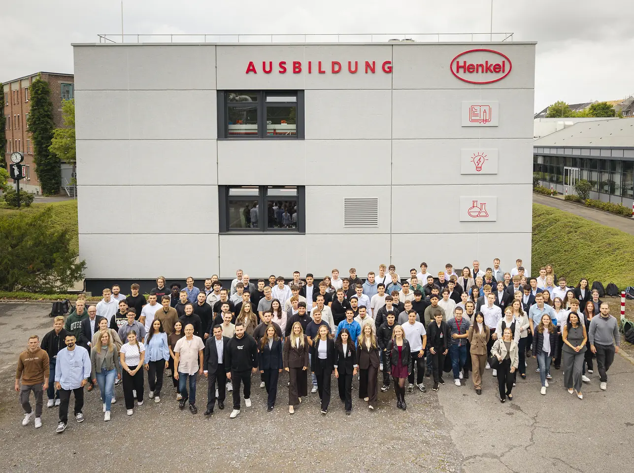 Eine große Gruppe neuer Auszubildender und dual Studierender steht vor dem Henkel-Ausbildungsgebäude in Düsseldorf zum Gruppenfoto. Über dem Gebäude ist das rote Henkel-Logo sowie die Aufschrift „Ausbildung“ zu sehen.