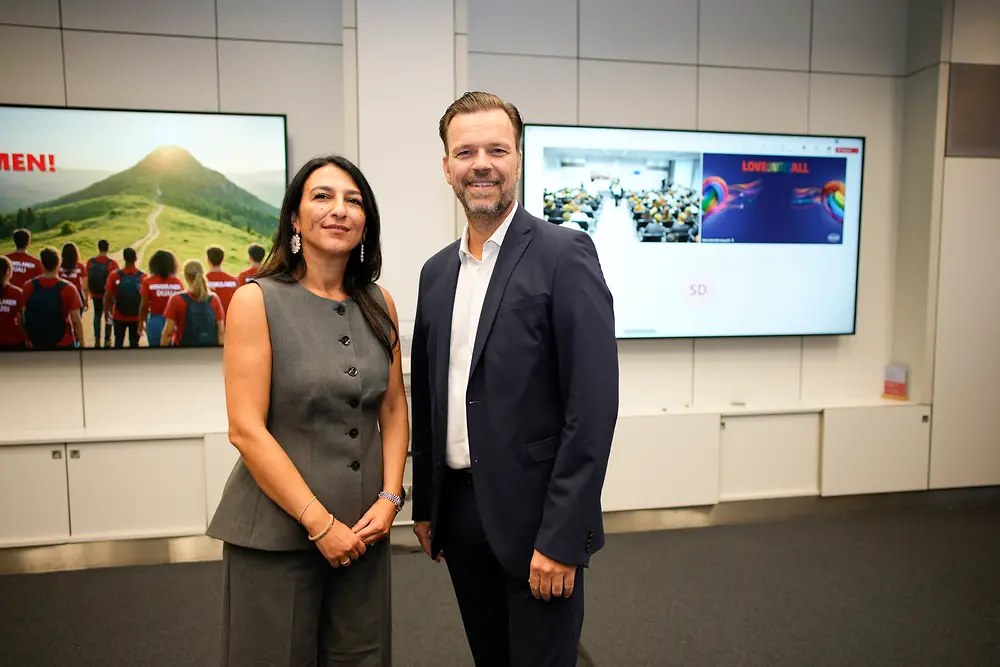 Henkel-Ausbildungsleiterin Funda Mutlu und Oliver Wilhelms, Personalleiter Deutschland und Schweiz, stehen nebeneinander und lächeln in die Kamera. Im Hintergrund sind Bildschirme mit Präsentationsfolien und Motiven zur Begrüßung der Auszubildenden zu sehen.