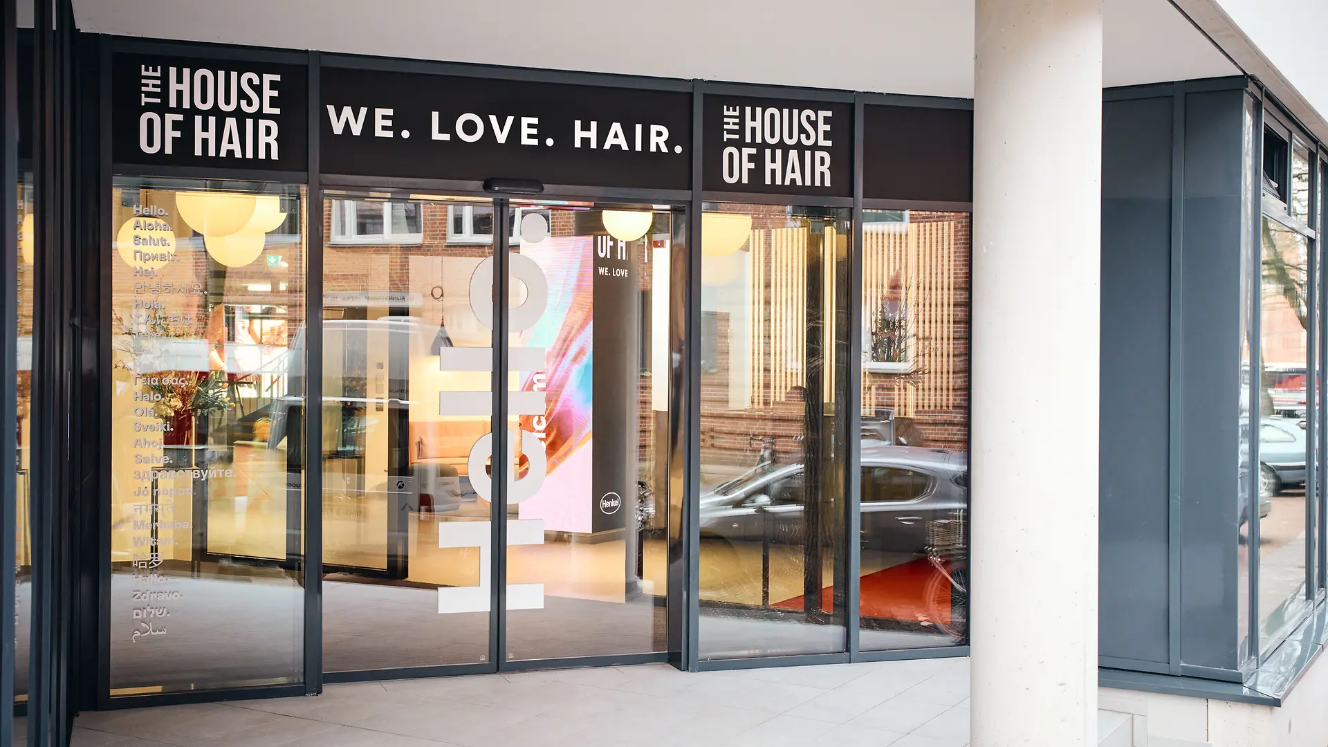 Verglaster Eingangangsbereich. Über der Tür die Slogan "We. Love. Hair". Rechts und links davon befindet sich jeweil die Aufschrift "The House of Hair".