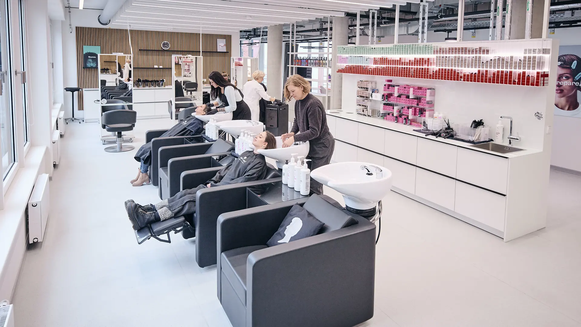 Friseursalon mit mehreren Haarwaschbecken. In zwei der Stühle sitzen Testkundinnen denen von zwei Mitarbeiterinnen die Haare gewaschen werden.