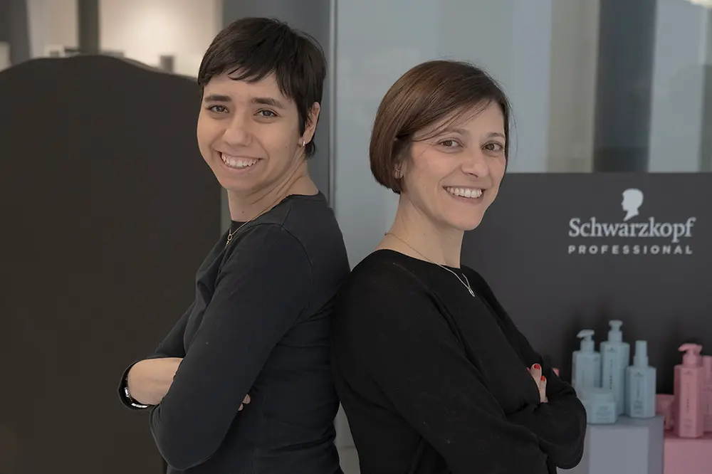 Noemí und ihre ehemalige Ausbilderin Meritxell stehen Rücken an Rücken im Salon und lachen seitlich in die Kamera.