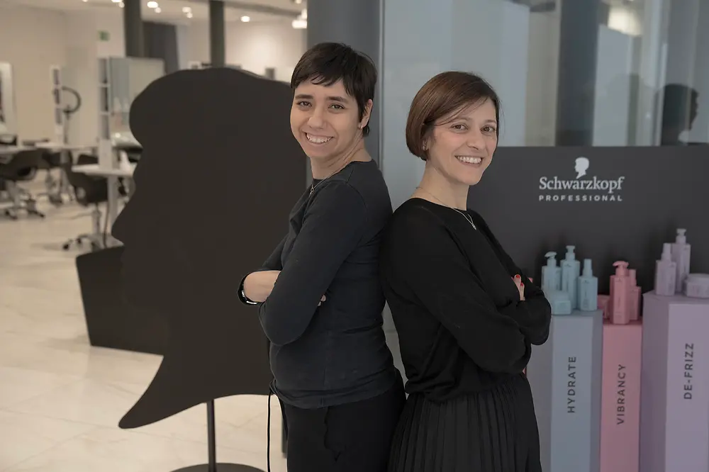 Noemí und ihre ehemalige Ausbilderin Meritxell stehen Rücken an Rücken im Salon und lachen seitlich in die Kamera.