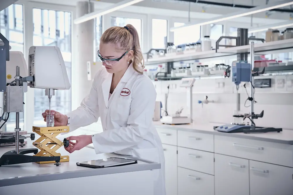 Eine Henkel-Mitarbeiterin aus der Forschung trägt einen weißen Laborkittel und Schutzbrille und arbeitet im Labor. Im Hintergrund sind Laborgeräte, Regale und große Fenster sichtbar.