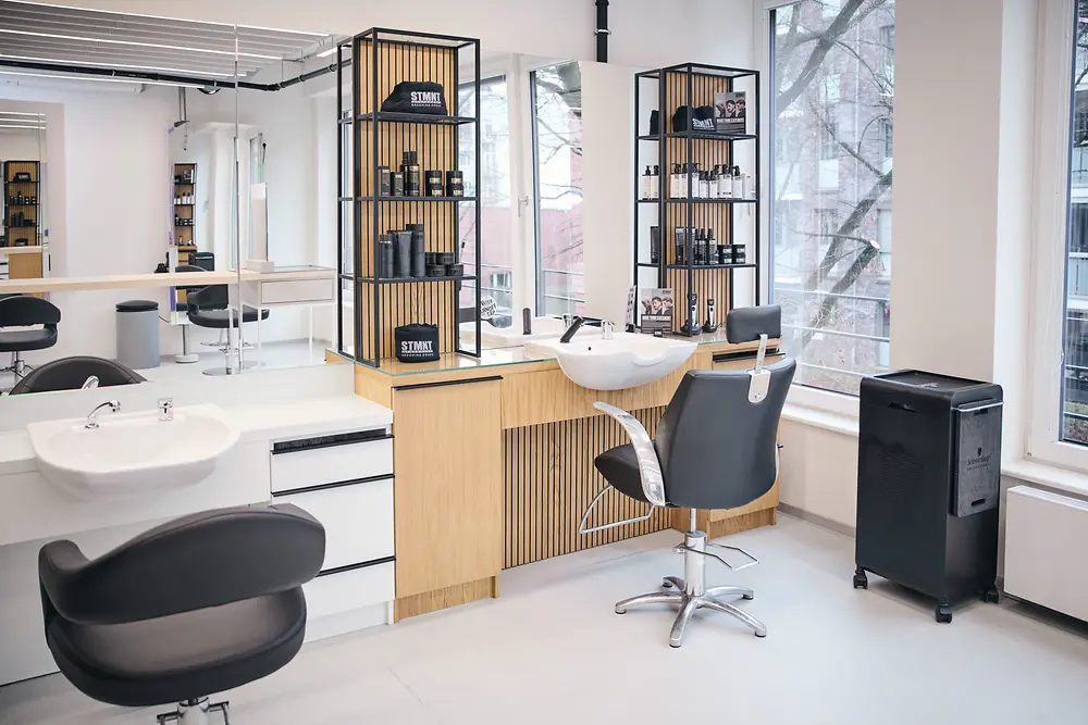 Heller, moderner Friseursalon mit zwei schwarzen Friseurstühlen vor Regalen mit Haarpflegeprodukten und zwei weißen Waschbecken. 