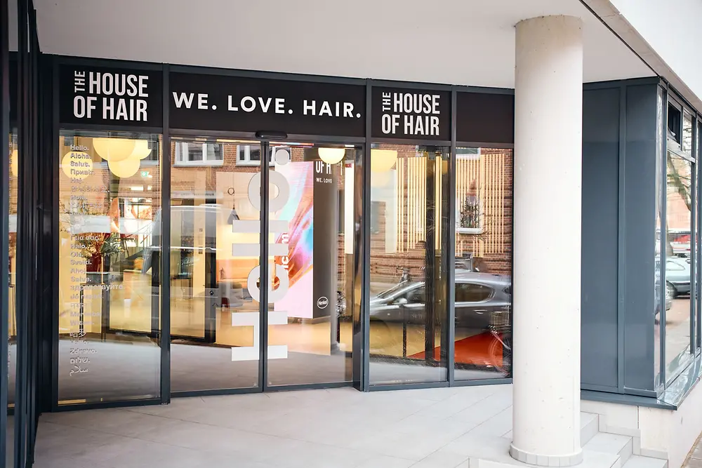 Eingangsbereich des House of Hair mit großen Glasfenstern und Türen. Über dem Eingang steht „WE. LOVE. HAIR.“.