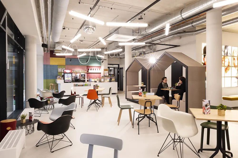 Heller Gemeinschaftsbereich im House of Hair mit vielfältigen Sitzmöglichkeiten, darunter zwei kleine Häuschen-Strukturen als private Sitzbereiche, in denen zwei Personen sitzen. Im Hintergrund befindet sich eine Theke mit Getränken und Snacks.