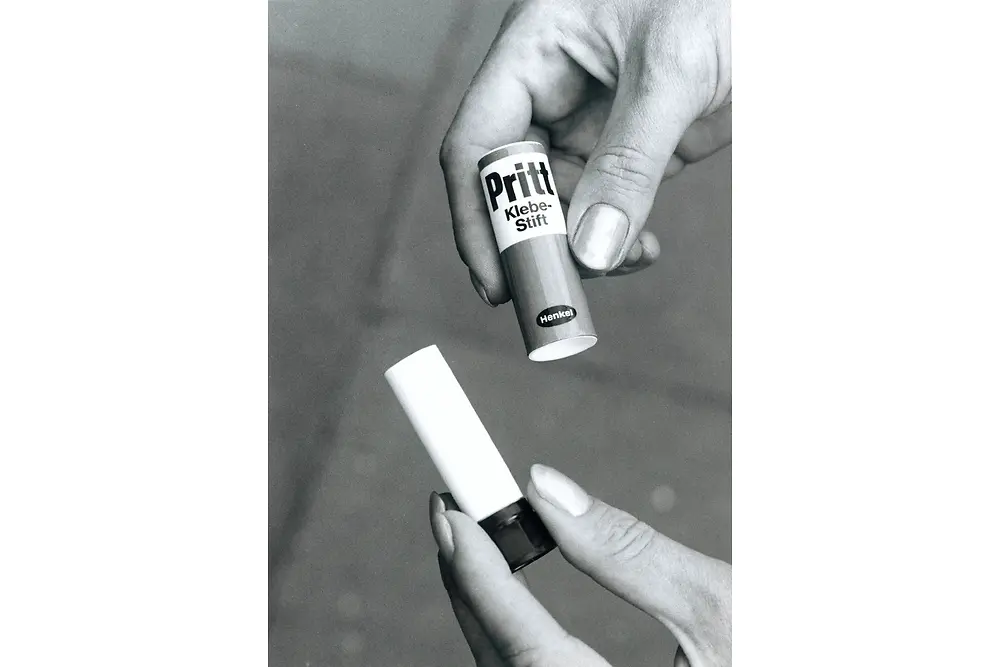 Schwarz-weiß-Foto: Zwei Hände halten einen Pritt-Klebestift. Eine Hand setzt die helle Kappe ab, die andere hält den Stift mit der Aufschrift „Pritt Klebe-Stift Henkel“
