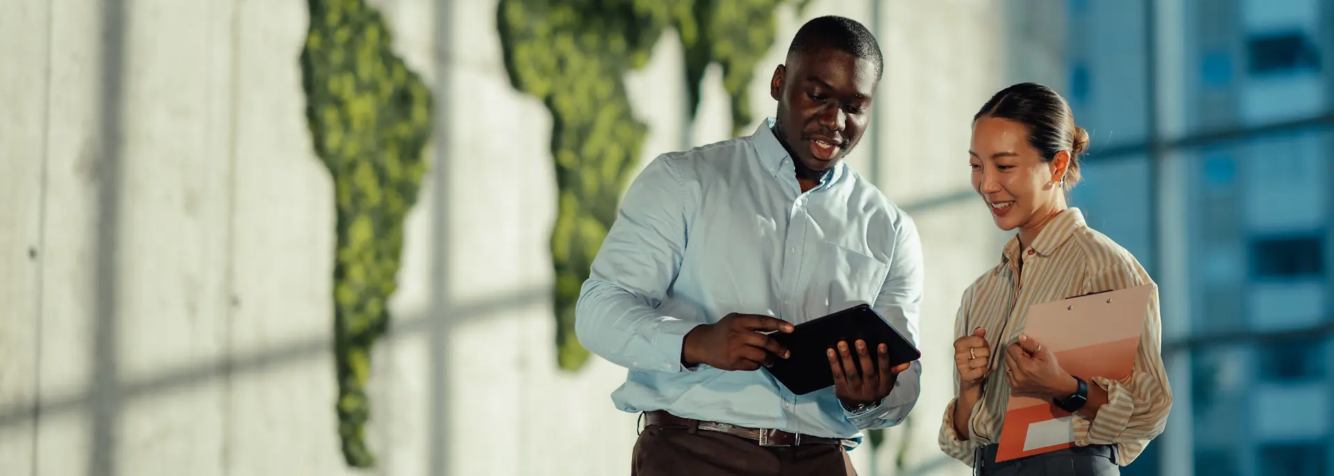 Two people standing in a modern office space, discussing information displayed on a tablet. A large green world map made from foliage decorates the wall behind them.