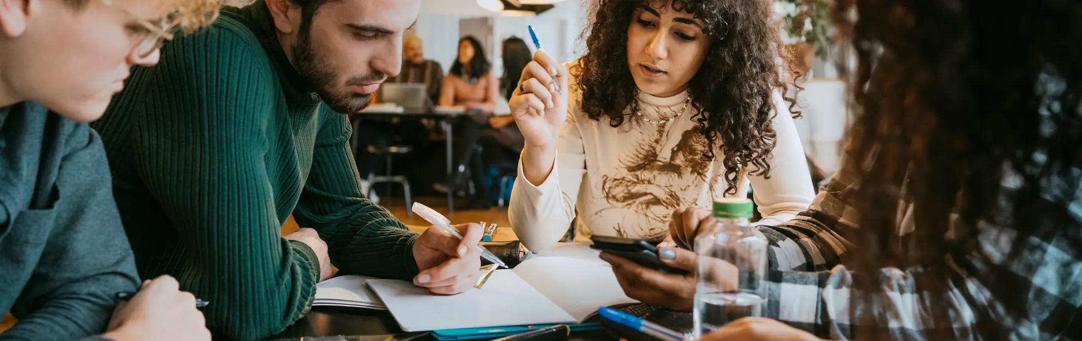 A group of people sitting together at a table in a modern indoor workspace, collaborating on documents, notebooks, and digital devices. Various pens, papers, and sticky notes are spread across the table while the group engages in discussion and note-taking.